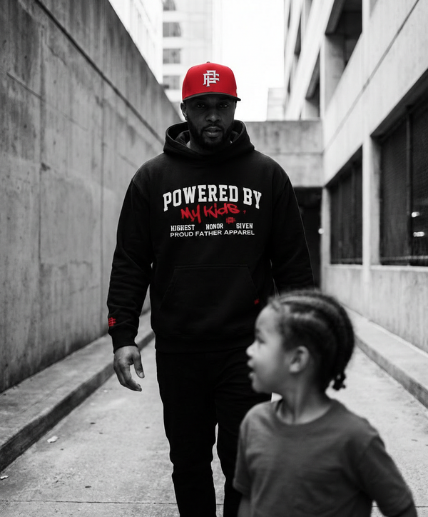 Man wearing a black hoodie with text and a red cap, walking with a child in an urban setting.