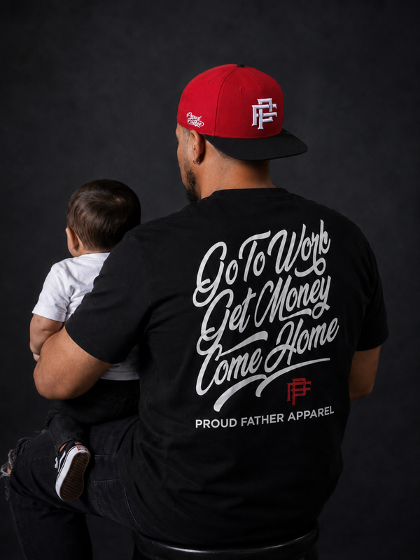 A man wearing a black t-shirt with a large white script graphic on the back and a red baseball cap sits holding a baby against a dark studio background. The t-shirt features the text 'Go To Work Get Money Home Come Home' and 'PROUD FATHER APPAREL'.